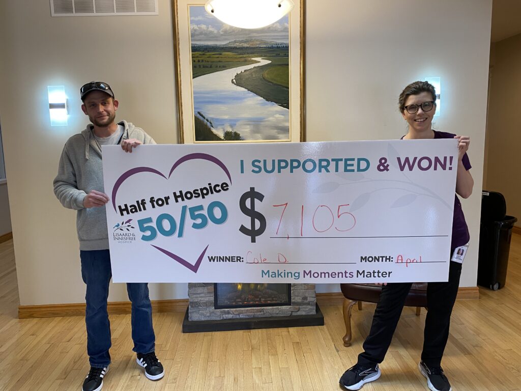 Two people stand indoors in front of a fireplace and landscape painting, holding an oversized cheque for the Lisaard & Innisfree Hospice 50/50 raffle. The cheque reads “Half for Hospice 50/50,” “I Supported & Won!,” and shows a prize amount of $7,105. The winner line reads “Cole D.” and the month is listed as April.