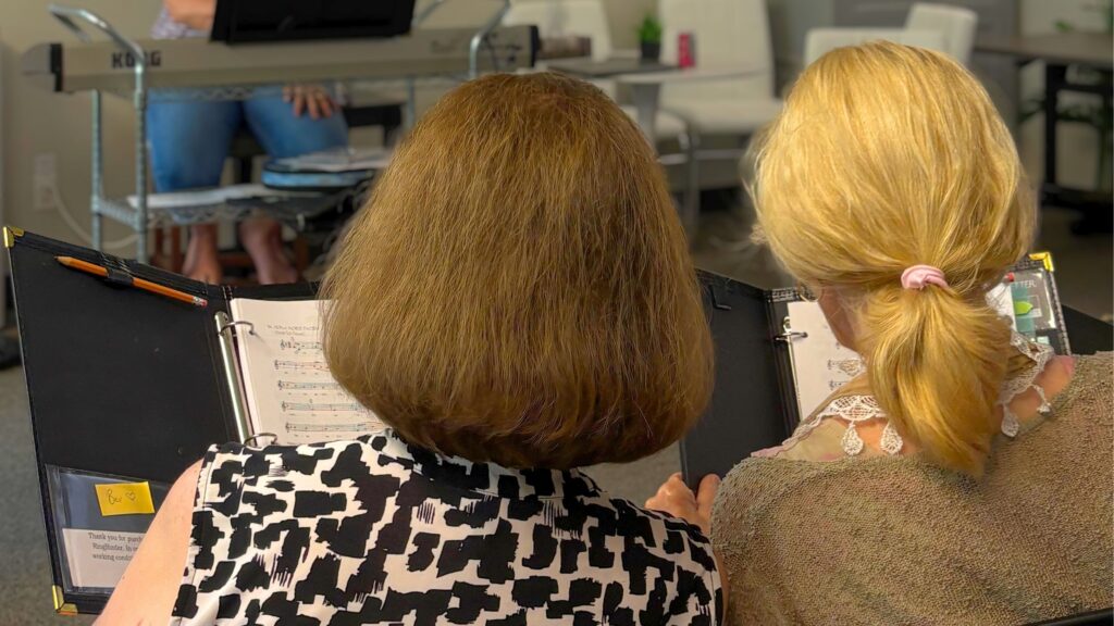 Image of the back of two individuals sitting side by side, holding choir books in front of them.